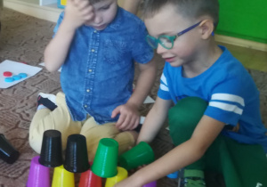 Dwóch chłopców buduje na dywanie wieżę z plastikowych kolorowych kubków